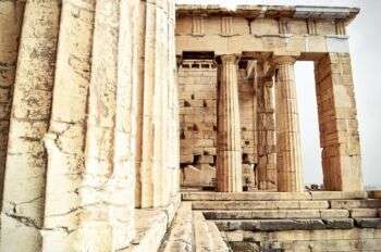 Gros plan du temple d’Athéna Nike. Il y a trois escaliers qui se tiennent devant trois colonnes du temple sur le côté droit. De plus, à gauche, il y a une colonne qui est très proche de la caméra. 