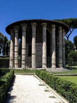 Temple de Vesta, à Rome. La photo a le temple au centre comme 8 des colonnes tenir le toit circulaire. Il y a un bain de gravier menant au temple en bas à gauche de l’écran. Une clôture en métal noir entoure le temple, un ciel bleu occupe le fond et des buissons verts entourent la structure. 