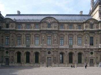 L’aile Lescot du Palais du Louvre, qui est un grand bâtiment brun en pierre avec une rangée d’arcs et deux rangées de fenêtres, la couche supérieure ayant des fenêtres plus petites que la rangée du milieu. Il y a aussi un toit sombre au-dessus du bâtiment. 