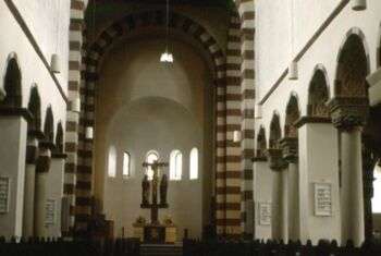 Hildesheim - Michaeliskirche Interior: A simple church with striped arches, Blank, tan-colored walls and a crucifix at the end of the isle. Small arched windows sit at on the middle of the dome near the back of the photo. 